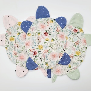 May include: Several handmade turtle-shaped fabric items. The turtle shells feature a floral pattern with pink, yellow, and red flowers on a white background. The flippers are pink, blue, and green with white polka dots. The turtles are arranged in a circular pattern.
