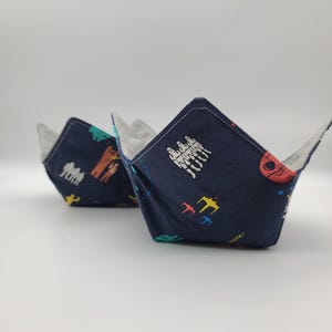May include: Two fabric bowl cozies featuring a dark navy blue exterior with a colorful repeating pattern of space-themed characters, starships, and red helmet icons. The interior of each bowl cozy is lined with a solid light gray fabric.
