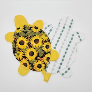 May include: Two turtle-shaped items. One has a black background with yellow sunflowers. The other is white with a green dotted pattern. Both have yellow flipper-like appendages and a yellow strap.