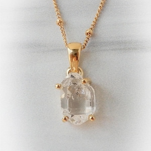 May include: A gold-colored necklace with a clear, faceted crystal pendant. The chain features small, round beads and a delicate link design. The pendant is an irregular, hexagonal shape with gold-colored accents.