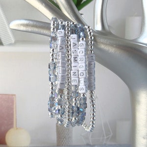 May include: A set of four beaded bracelets featuring silver-toned metallic beads and light blue faceted crystal beads. The bracelets include white square letter beads spelling out the words GRATEFUL, JASMINE, GORGEOUS, and DAVID, displayed on a silver hand.
