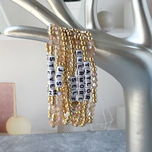 Peut inclure: Un ensemble de bracelets en perles dorées avec des perles de lettres blanches épellant des noms, dont "Taylor", "Vermore", "Self", "Juls" et "Steven".