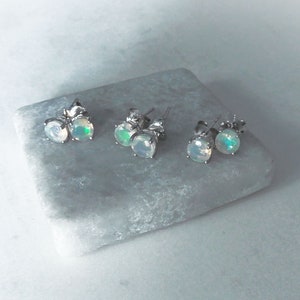 May include: Three pairs of silver stud earrings with iridescent opal stones. The earrings are displayed on a light gray stone surface.