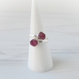 May include: A silver ring with two pink gemstones set in the band. The ring is displayed on a white ring stand.