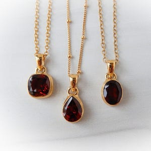 May include: Three gold necklaces with pendants featuring dark red gemstones. The pendants have different shapes: a square, a teardrop, and an oval. The chains are delicate and the gemstones are set in gold.