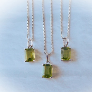 May include: Three silver necklaces with rectangular, light green pendants. The pendants are a vibrant, olive green color. The chains are delicate and hang against a white background. The necklaces are displayed in a vertical arrangement.