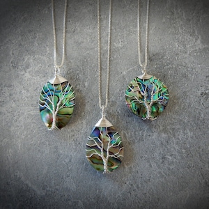 May include: Three silver necklaces with abalone shell tree of life pendants. The pendants are oval, round, and teardrop shaped. The abalone shell is iridescent with shades of green, blue, and purple.