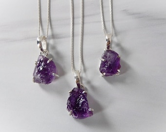 May include: Three silver necklaces with raw amethyst pendants. The amethyst stones are a deep purple color and have a rough, natural texture.