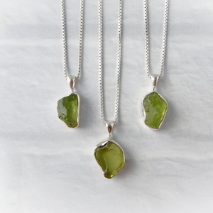 May include: Three silver necklaces with olive-green gemstone pendants. Each pendant has a unique, irregular shape and is set in a silver bezel. The necklaces hang against a white and gray background.