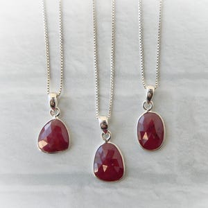 May include: Three silver necklaces with ruby pendants. Each pendant features a faceted, deep red gemstone in a different shape: a rounded triangle, a teardrop, and an oval. The necklaces hang against a light gray background.