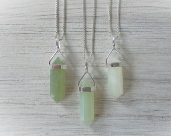May include: Three silver necklaces with green jade crystal point pendants hanging against a white wood background.