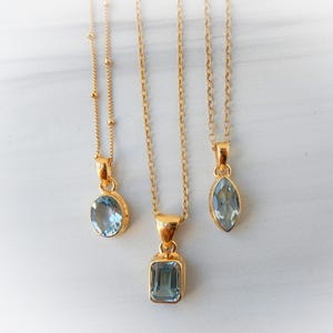 Może przedstawiać: Trzy złote naszyjniki z wisiorkami z jasnoniebieskich kamieni szlachetnych. Wisiorki mają różne kształty: owalny, prostokątny i markiza. Naszyjniki są prezentowane na białej marmurowej powierzchni.