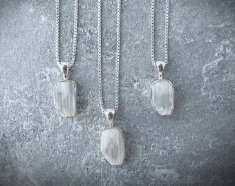 Puede incluir: Tres collares de plata con colgantes de selenita blanca cruda y translúcida. Cada colgante está sujeto por una reasa de plata pulida y cuelga de una delicada cadena de plata. Las piedras presentan una textura fibrosa, vertical y formas.