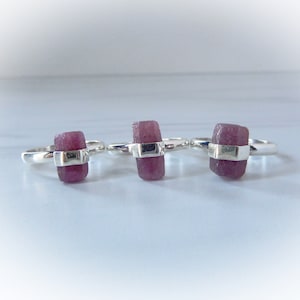 May include: Three silver rings each featuring a raw, rectangular, pink gemstone. The gemstones are held in place by a silver band. The rings are displayed on a white surface.