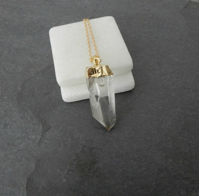 Puede incluir: Un collar de cadena dorada con un colgante de cristal de cuarzo transparente. El colgante est&aacute; engastado en un bisel dorado y tiene una punta puntiaguda.