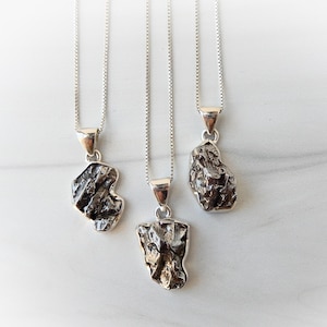 May include: Three silver necklaces with unique, irregularly shaped pendants. The pendants have a textured, dark gray appearance, and are suspended from delicate silver chains. The necklaces are displayed against a white marble background.