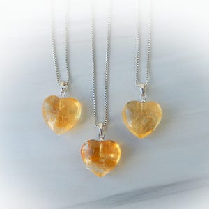 May include: Three necklaces featuring heart-shaped yellow citrine pendants suspended from thin silver box chains. Each pendant is a polished, translucent gemstone with a warm golden hue, attached to the chain by a small silver bail.