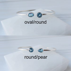 May include: Two silver bangle bracelets with blue gemstones. The top bracelet has an oval and round gemstone. The bottom bracelet has a round and pear-shaped gemstone. The text "oval/round" and "round/pear" is below each bracelet.