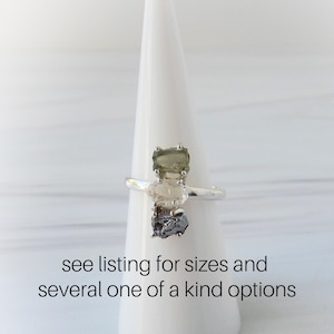 May include: A silver ring with three raw, rectangular gemstones in shades of green, clear, and dark gray. The ring is displayed on a white cone-shaped stand. Text on the image reads: "see listing for sizes and several one of a kind options."