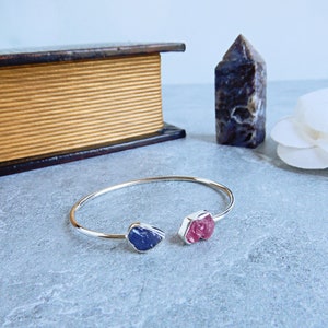 May include: A silver open cuff bracelet with a raw, natural gemstone at each end. One gemstone is a deep blue, the other is a vibrant pink. The bracelet is displayed on a gray surface, with a book and a crystal in the background.