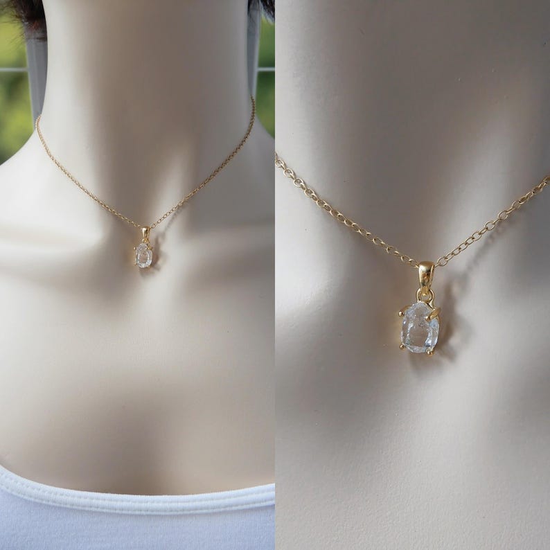 May include: A gold-coloured necklace featuring a clear, oval-shaped pendant. The pendant is set in a gold-coloured frame and suspended from a delicate gold chain. The necklace is displayed on a mannequin.
