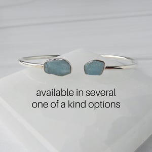 May include: A silver bangle bracelet with two light blue aquamarine stones. The bracelet is available in several one-of-a-kind options.