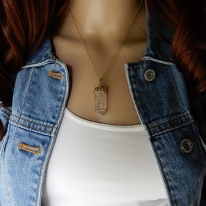 Puede incluir: Un collar de cadena dorada con un colgante de cristal de cuarzo transparente. El colgante est&aacute; facetado y tiene un engaste dorado.