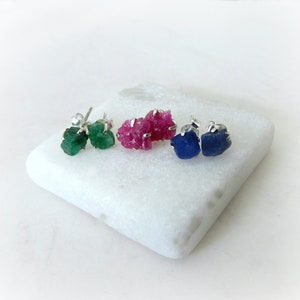 May include: Three pairs of raw gemstone earrings. The earrings are made with emerald, ruby, and sapphire gemstones. The gemstones are set in silver.
