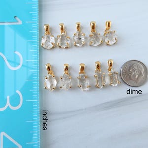 May include: A collection of gold-coloured pendants with clear, rough-cut crystal stones. The pendants are displayed on a white surface next to a dime and a ruler marked in inches. The pendants vary in shape and size.