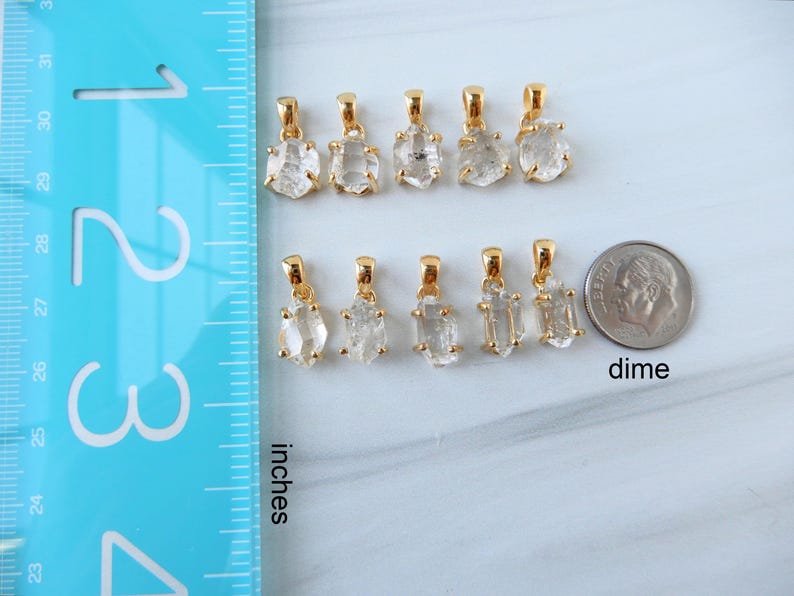 May include: A collection of gold-coloured pendants with clear, rough-cut crystal stones. The pendants are displayed on a white surface next to a dime and a ruler marked in inches. The pendants vary in shape and size.