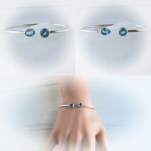 May include: A silver bangle bracelet with two blue topaz gemstones. The bracelet is shown on a mannequin's wrist and on a white background.