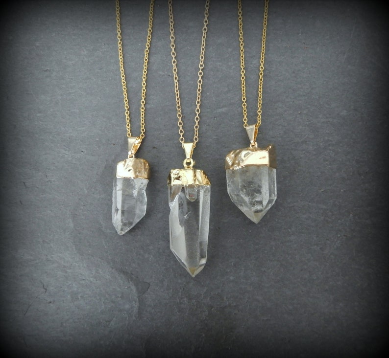 Puede incluir: Tres collares de oro con colgantes de cristal de cuarzo transparente. Los colgantes son de diferentes tama&ntilde;os y formas. Los collares est&aacute;n sobre un fondo gris.