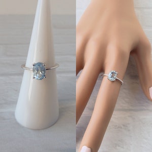May include: A silver ring with a light blue oval gemstone. The ring is on a white ring display stand and on a model's finger.