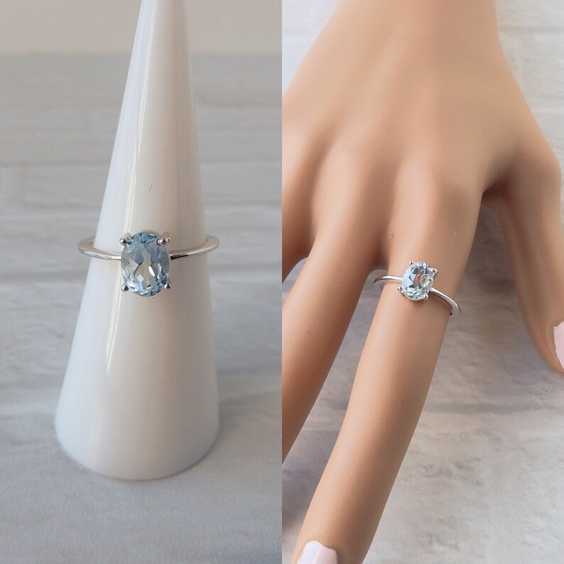 May include: A silver ring with a light blue oval gemstone. The ring is on a white ring display stand and on a model's finger.
