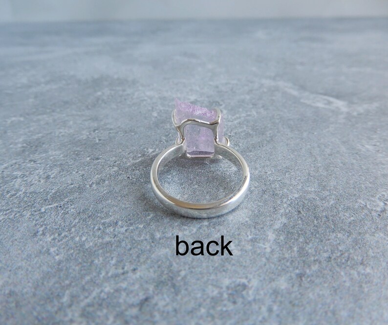 K&ouml;nnte beinhalten: Ein silberner Ring mit einem rosafarbenen Kristall in der Mitte. Die R&uuml;ckseite des Rings ist auf dem Bild zu sehen. Der Ring liegt auf einer grauen Oberfl&auml;che. Das Wort "back" steht unter dem Ring.