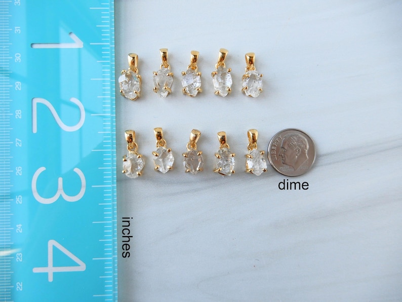 May include: Ten gold pendants with clear, irregular-shaped stones are shown. A dime is next to the pendants for size comparison. A blue ruler with inch markings is on the left side of the image.