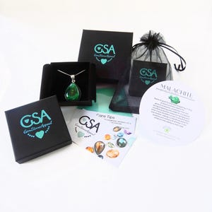 May include: A green malachite gemstone pendant necklace in a black box with a white background. The necklace is packaged with a black velvet bag and a card with information about the stone. The card reads "Malachite, prosperity, protection, renewal, abundance".