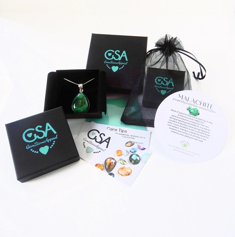 May include: A green malachite gemstone pendant necklace in a black box with a white background. The necklace is packaged with a black velvet bag and a card with information about the stone. The card reads "Malachite, prosperity, protection, renewal, abundance".
