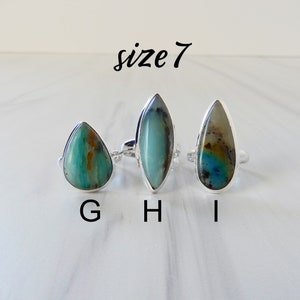 May include: Three silver rings with different green and blue gemstone settings. The rings are labeled G, H, and I. The text "size 7" is above the rings.