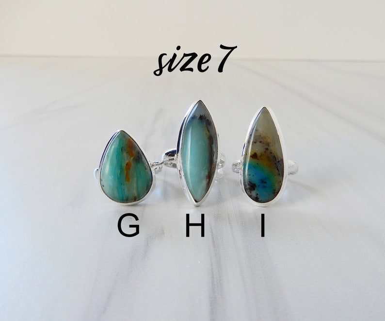 May include: Three silver rings with different green and blue gemstone settings. The rings are labeled G, H, and I. The text "size 7" is above the rings.