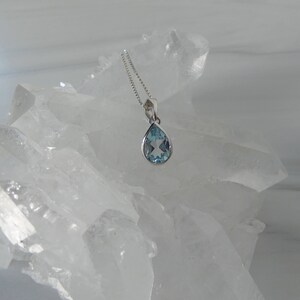May include: A teardrop-shaped blue topaz pendant necklace on a silver chain. The pendant is set in a silver bezel and is surrounded by clear quartz crystals.