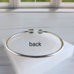 May include: A silver bangle bracelet with two blue gemstones. The bracelet is on a white surface and the word "back" is written below the bracelet.