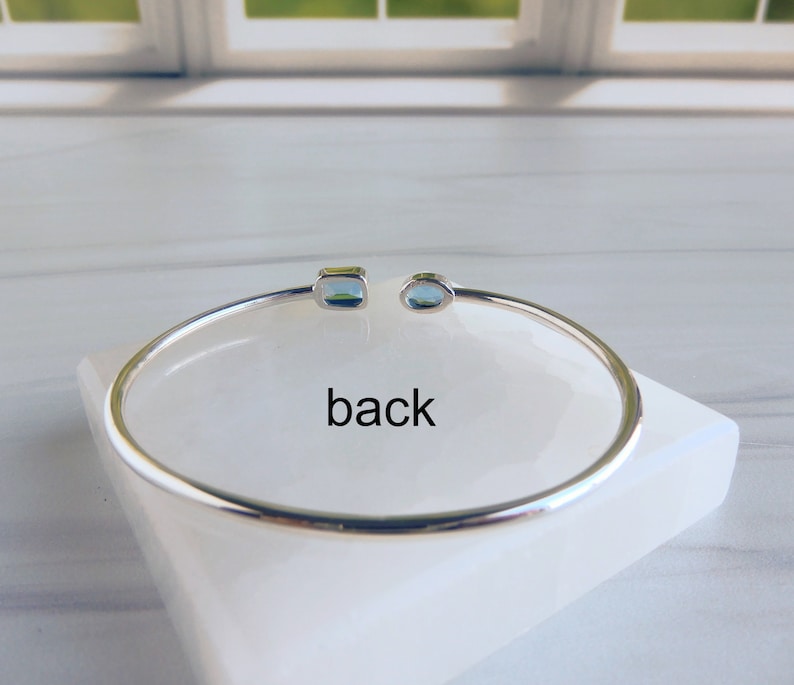 May include: A silver bangle bracelet with two blue gemstones. The bracelet is on a white surface and the word "back" is written below the bracelet.