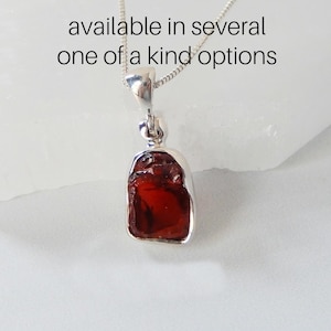 May include: A silver chain necklace with a pendant featuring a raw, red garnet gemstone. The pendant is set in a silver bezel and has a simple design. The necklace is available in several one-of-a-kind options.