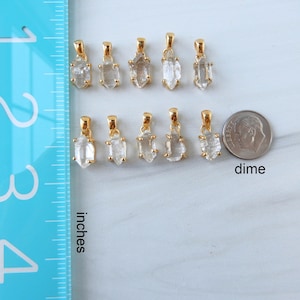 May include: A selection of gold-plated crystal pendants displayed on a white surface. A blue ruler marked in inches and a dime are placed nearby for scale. The pendants are made of clear, raw crystal stones. The word "inches" is printed on the ruler.
