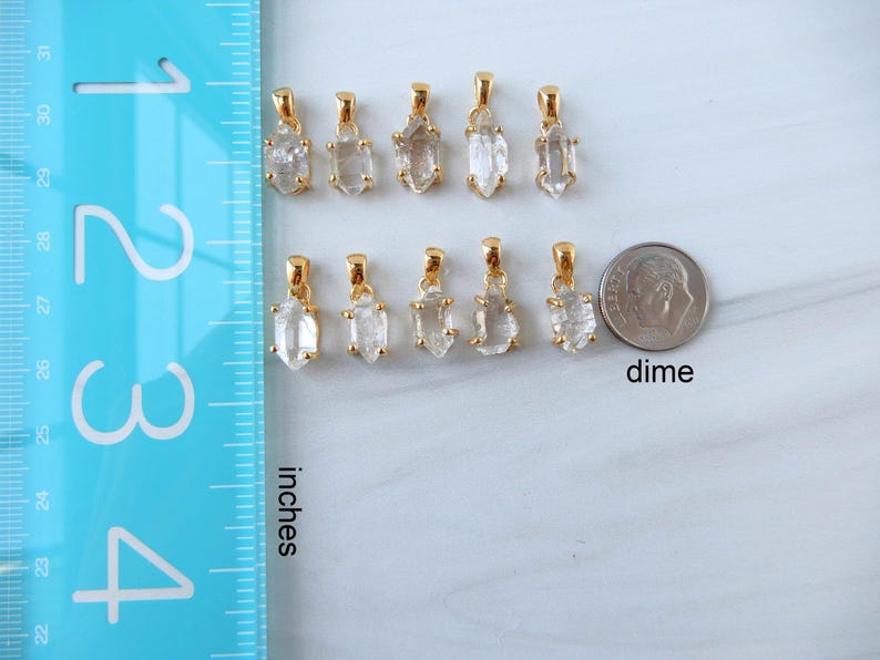 May include: A selection of gold-plated crystal pendants displayed on a white surface. A blue ruler marked in inches and a dime are placed nearby for scale. The pendants are made of clear, raw crystal stones. The word "inches" is printed on the ruler.