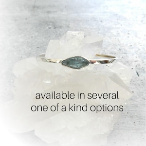 May include: A silver bangle bracelet with a light blue gemstone. The bracelet is available in several one-of-a-kind options.