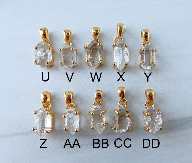 May include: Ten gold-coloured pendants featuring clear, raw crystal stones. Each pendant has a unique shape and is labelled with a letter from U to DD. The pendants are displayed on a light-coloured surface.