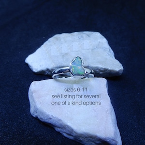 May include: A silver ring with a light green opal gemstone. The ring is set on a white rock. The text "sizes 6-11 see listing for several one of a kind options" is written below the ring.