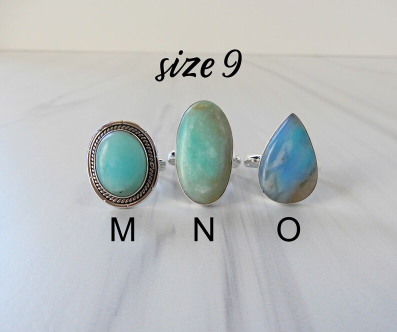 May include: Three silver rings with different colored gemstones. The ring on the left has a round, light blue gemstone with a silver border. The middle ring has an oval, light green gemstone. The ring on the right has a teardrop-shaped, light blue gemstone. The rings are labeled M, N, and O, respectively. The text "size 9" is above the rings.
